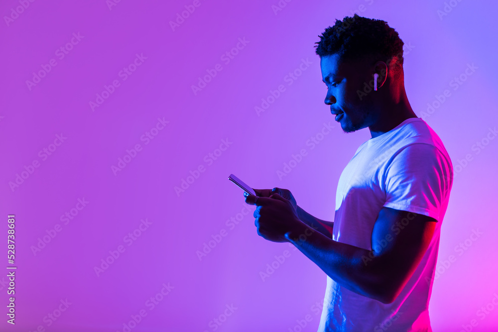 Side view of young black man in earphones listening to music, using new mobile app on smartphone in neon light, free space - obrazy, fototapety, plakaty 