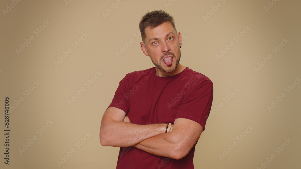 Cheerful funny bully young man in red t-shirt. Adult guy showing tongue making faces at camera, fooling around, joking, aping with silly face, teasing isolated alone on beige studio background indoors Cheerful funny bully young man in red t-shirt. Adult guy showing tongue making faces at camera, fooling around, joking, aping with silly face, teasing isolated alone on beige studio background indoors