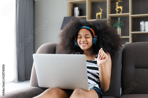 A cute little girl wearing headphones is sitting on her laptop on the sofa on her vacation. photo