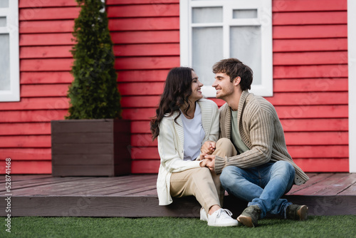 Side view of young couple in warm cardigans holding hands near blurred house