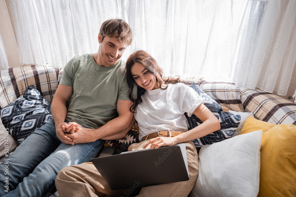 Smiling man holding hand of girlfriend using laptop on bed in camper