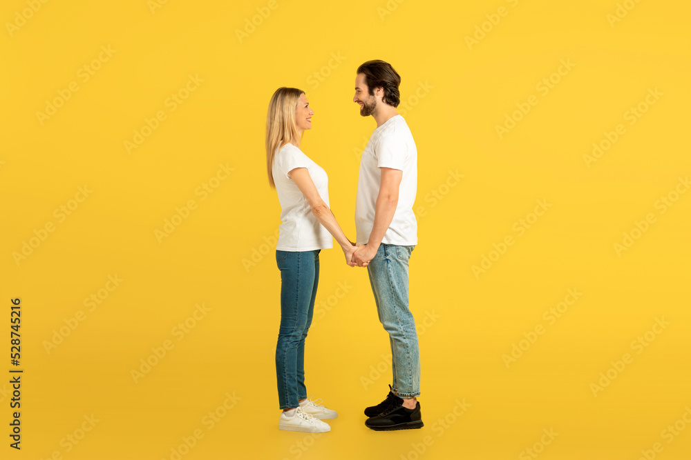 Cheerful millennial caucasian male and female in white t-shirts holding hands, looking at each other Cheerful millennial caucasian male and female in white t-shirts holding hands, looking at each other
