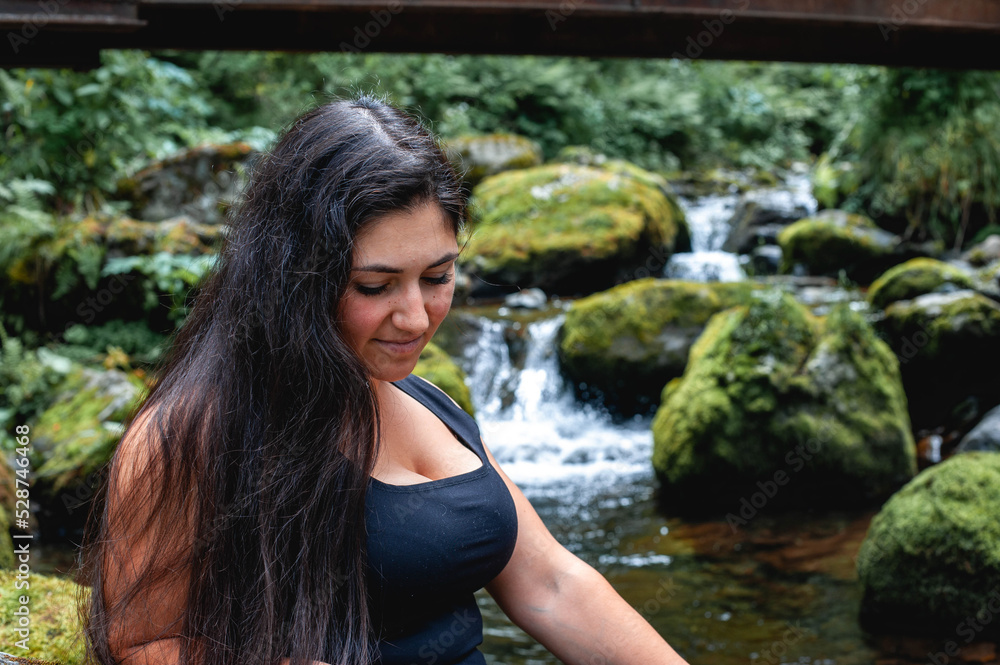 a young modest Turkish woman with black and long hair, a beautiful face and large breasts in a T-shirt sits by a mountain stream among large stones covered with green moss and looks down
