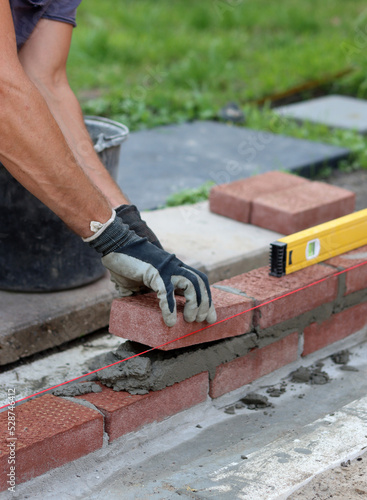Male hands in protective gloves holding red bricks. Construction worker builds masonry. Brickwork close up photo. 