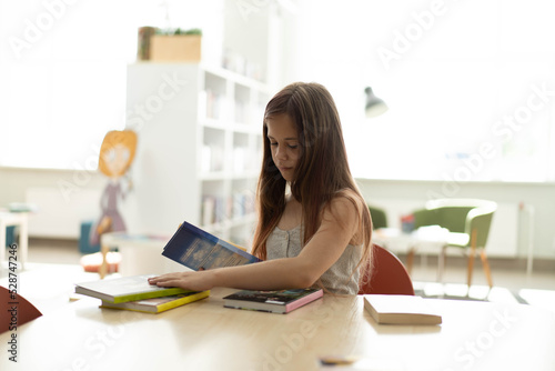  A teenage girl among piles of books. A young caucasian girl is reading a book against the background of shelves. She is surrounded by stacks of books. Book Day.