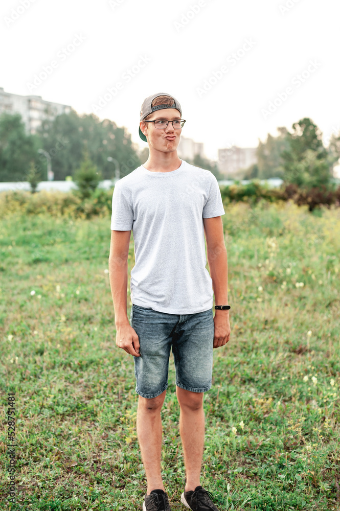 Fototapeta premium A cheerful Caucasian guy with glasses and a cap on his head is dressed in a white T-shirt and jeans made a grimace on his face, stretching his lips. Summer photo at sunset