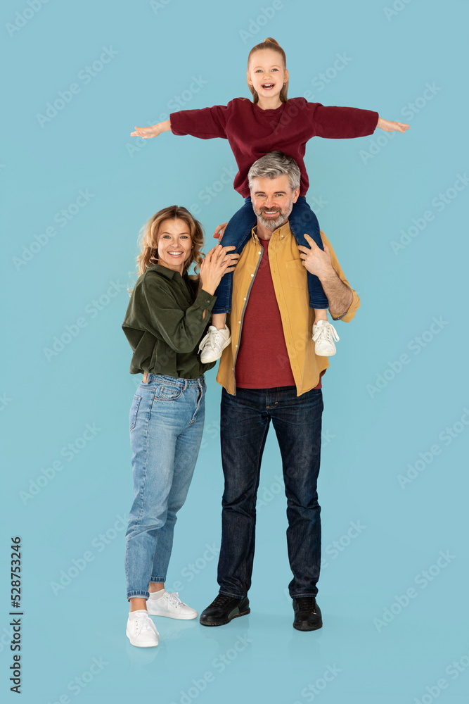 Mother And Father Posing, Holding Daughter On Shoulders, Blue Background Mother And Father Posing, Holding Daughter On Shoulders, Blue Background