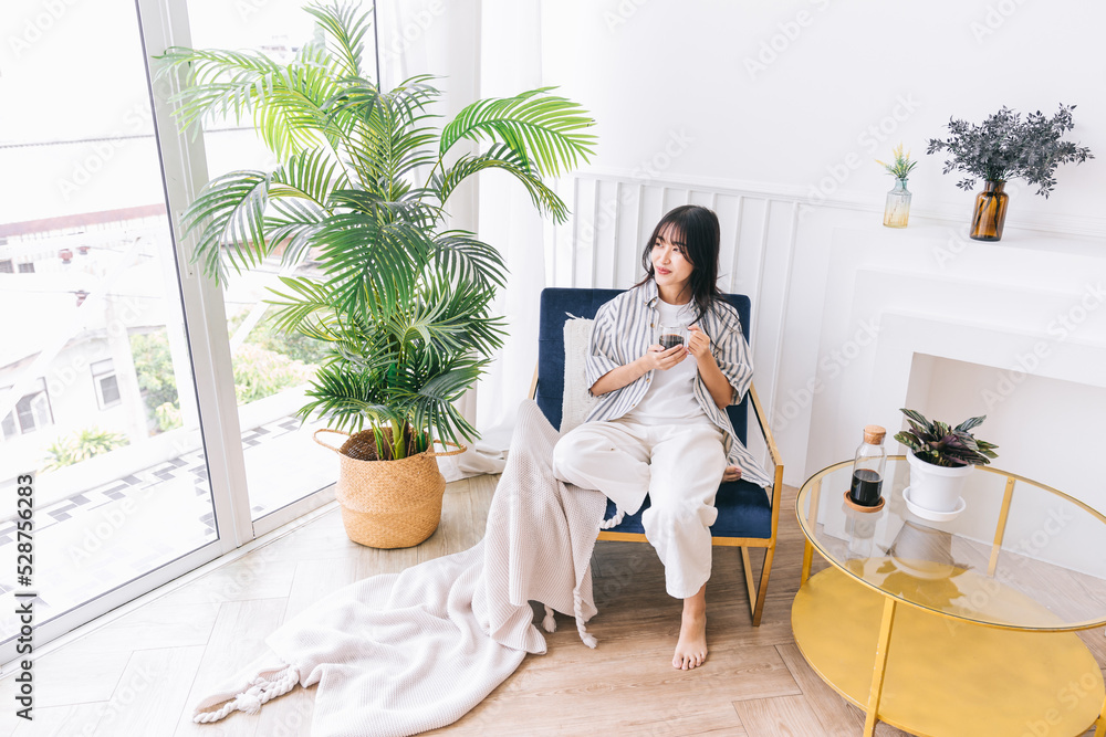 Young pretty woman smile and sits in the chair of the modern living area with a green plant, Relaxing drinking a cup of hot coffee, Healing reflection in an indoor environment at a cozy home. Young pretty woman smile and sits in the chair of the modern living area with a green plant, Relaxing drinking a cup of hot coffee, Healing reflection in an indoor environment at a cozy home.