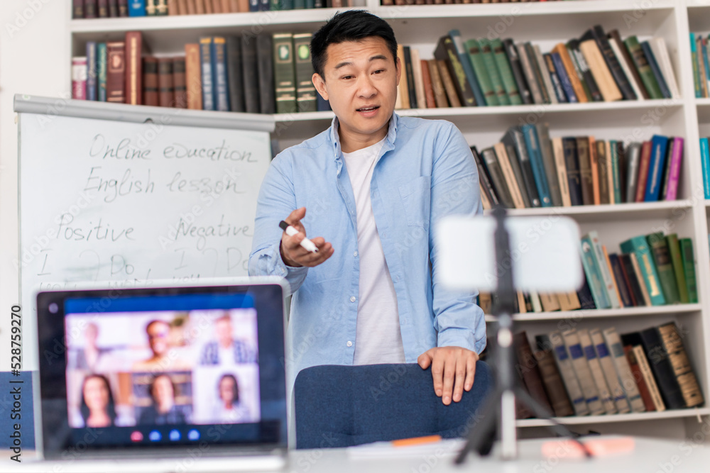 Asian Tutor Talking To Laptop And Cellphone Having Lecture Indoor Asian Tutor Talking To Laptop And Cellphone Having Lecture Indoor