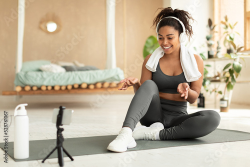 Fitness blogging concept. Fit black lady shooting online trainings using smartphone on tripod, sitting on yoga mat