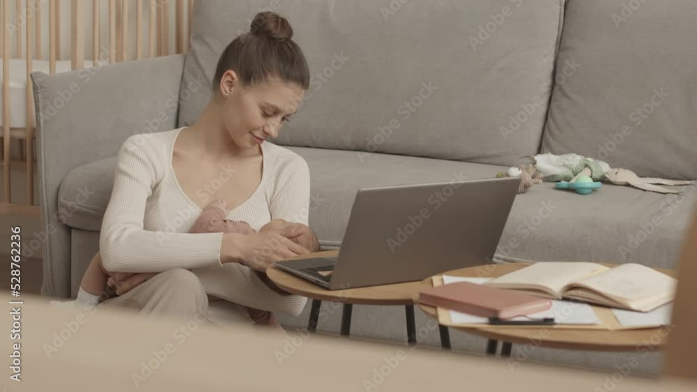Medium slowmo of young Caucasian woman on maternity leave working or studying on laptop from home with newborn baby daughter sleeping in her arms Medium slowmo of young Caucasian woman on maternity leave working or studying on laptop from home with newborn baby daughter sleeping in her arms