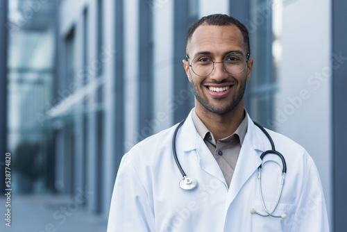 Close-up photo portrait of young African American doctor outside hospital  man in medical coat smiling and looking at camera.