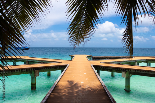 Fototapeta Naklejka Na Ścianę i Meble -  Wooden walkways on the pier of a resort island with sand marks in the Maldives against the background of hanging palm leaves, a sky with clouds and clear turquoise water of the ocean and a boat