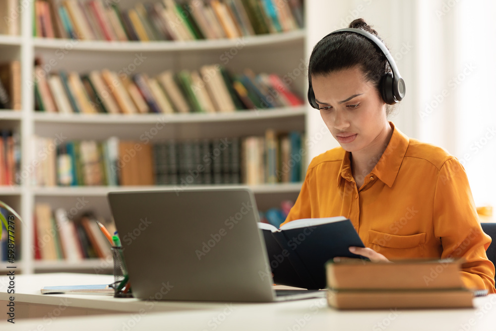 Focused female student reading book and using laptop computer, studying online, sitting in library, copy space Focused female student reading book and using laptop computer, studying online, sitting in library, copy space