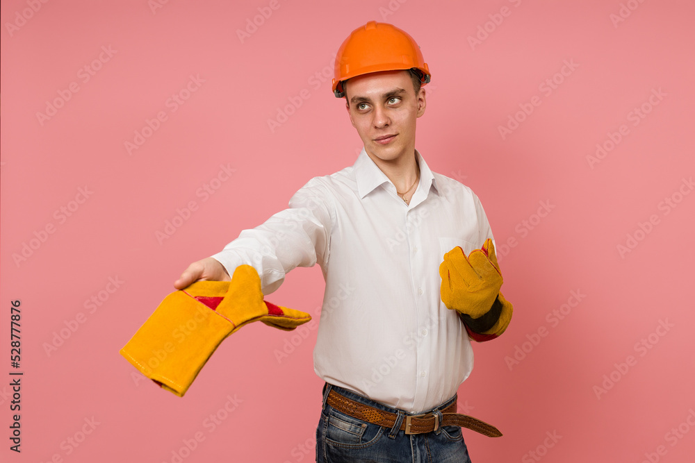 a young man in a white shirt and hard hat gives protective gloves and stands against a pink background