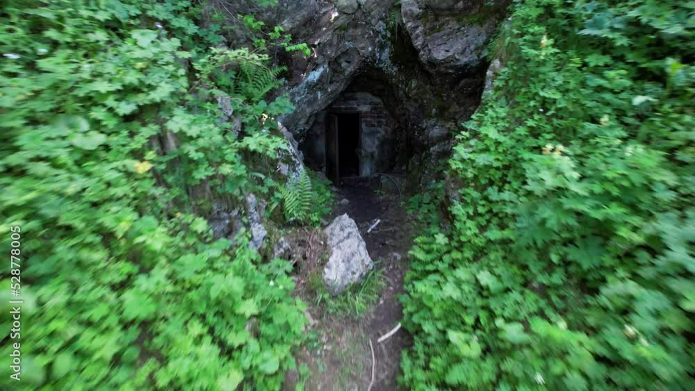 Hidden entrance to the cave among green grass and bushes. Mountainous terrain. The path leads to a cave with an old door. Steep cliffs. Coniferous forest. A fern is growing. Mountains of Kazakhstan Hidden entrance to the cave among green grass and bushes. Mountainous terrain. The path leads to a cave with an old door. Steep cliffs. Coniferous forest. A fern is growing. Mountains of Kazakhstan