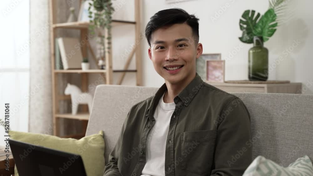 half length portrait of an attractive asian young man turning to smile at the camera while working from home on the computer in a cozy bright living room. half length portrait of an attractive asian young man turning to smile at the camera while working from home on the computer in a cozy bright living room.