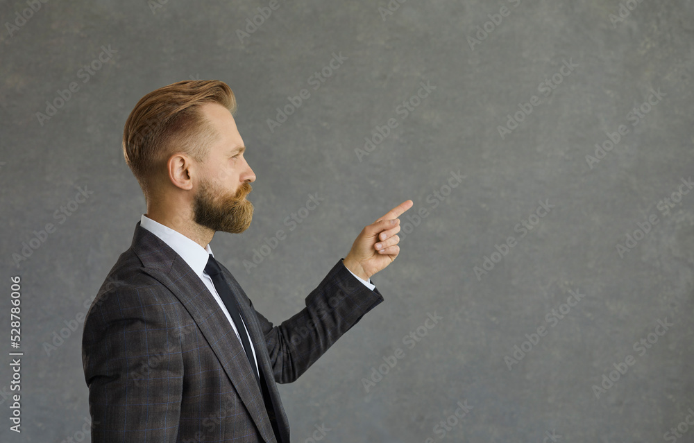 Profile portrait of young businessman advertising business idea. Side view of handsome man in smart suit looking and pointing finger aside at free text copy space standing isolated on grey background Profile portrait of young businessman advertising business idea. Side view of handsome man in smart suit looking and pointing finger aside at free text copy space standing isolated on grey background