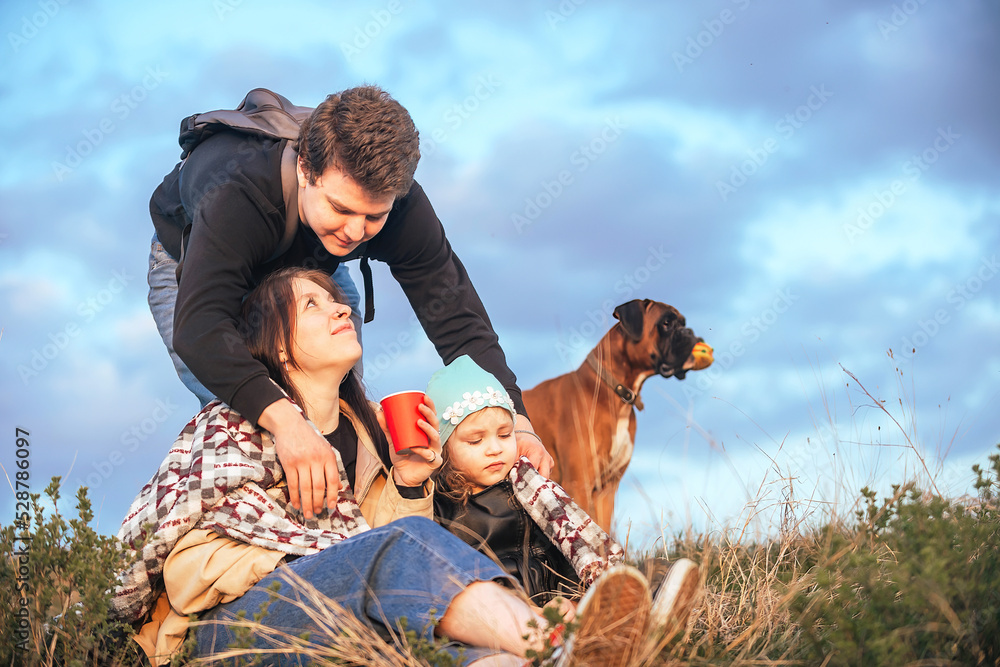 Fototapeta premium happy family of travelers, a man covers with a blanket and hugs his wife and daughter in nature outside the city against the backdrop of a bright sunset sky with clouds, their big dog stands behind