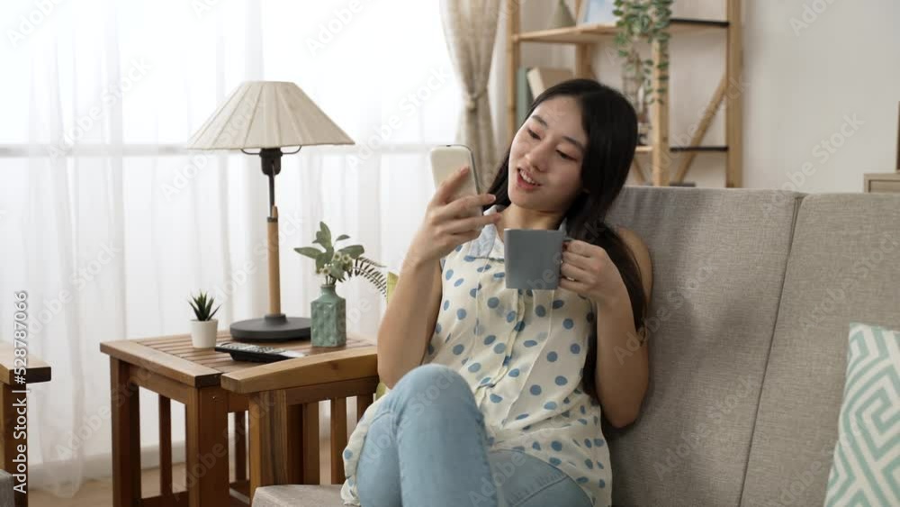 leisure Asian young female sitting down on living room couch carrying cup and phone is drinking coffee while reading fun online message at a bright home interior leisure Asian young female sitting down on living room couch carrying cup and phone is drinking coffee while reading fun online message at a bright home interior