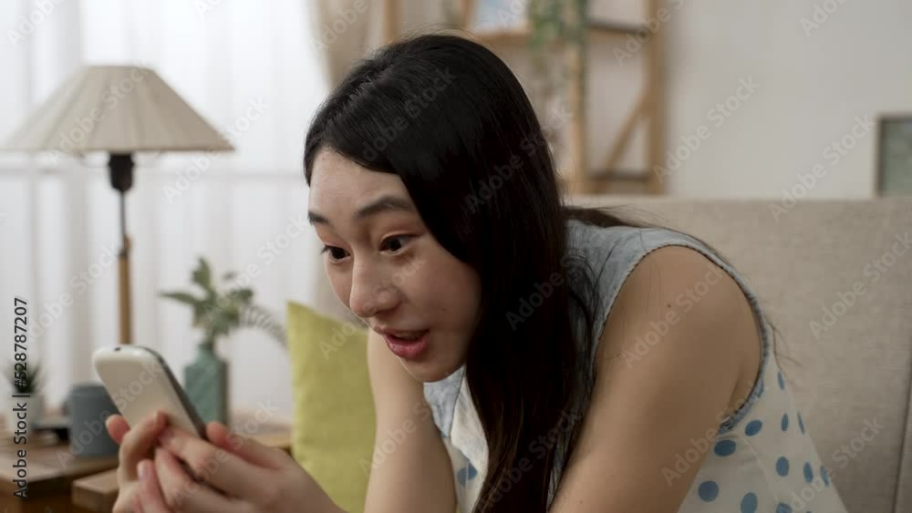 closeup of an excited Asian female talking to her self in surprised as she receives an online discount news on the mobile phone in the living room at home. closeup of an excited Asian female talking to her self in surprised as she receives an online discount news on the mobile phone in the living room at home.