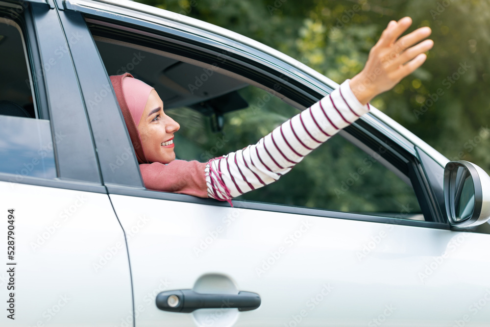 Glad cheerful young arab woman in hijab enjoy traveling on new car touches air with her hand through open window