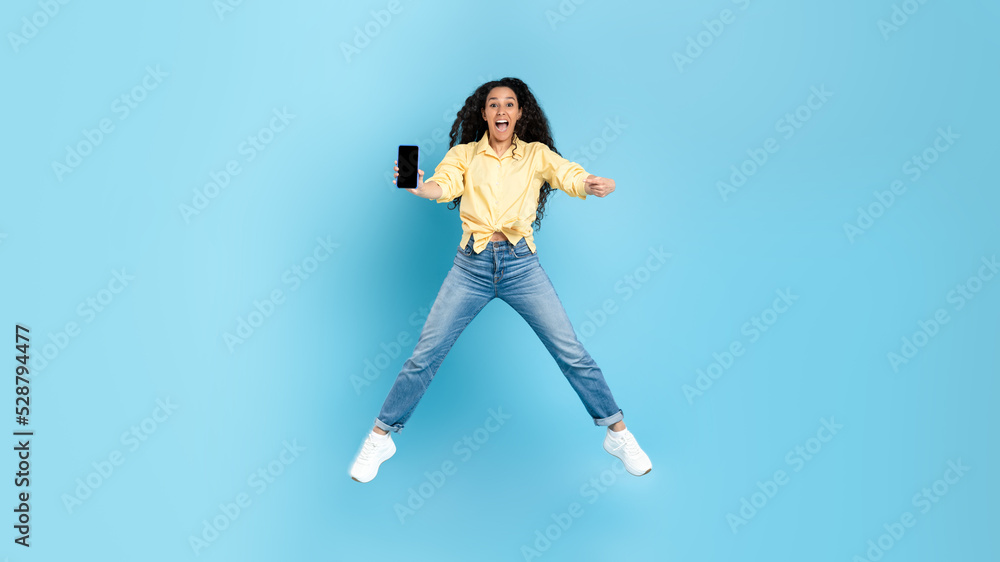 Excited Middle Eastern Woman Showing Phone Jumping Over Blue Background