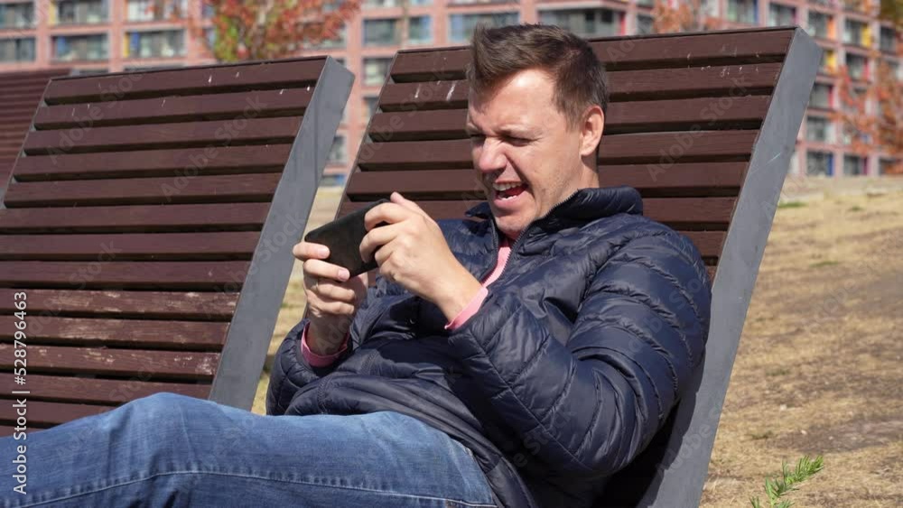 Excited man actively plays virtual games on his phone, frantically gestures with his hands and celebrates winning on a wooden chaise longue in the park in autumn Excited man actively plays virtual games on his phone, frantically gestures with his hands and celebrates winning on a wooden chaise longue in the park in autumn