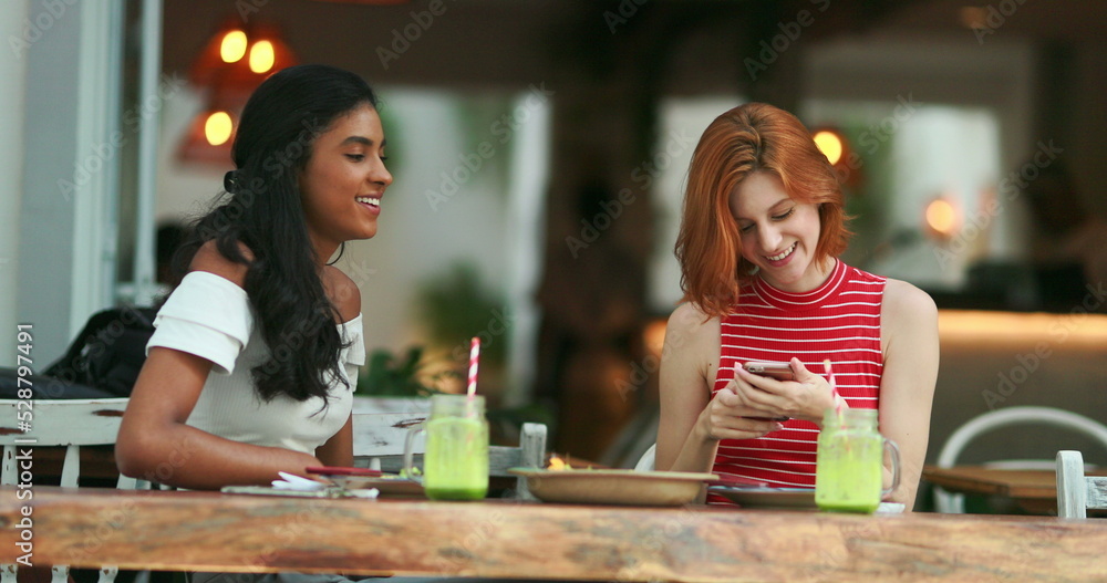 Two multiracial friends at cafe showing cellphone screen smiling and feeling happy Two multiracial friends at cafe showing cellphone screen smiling and feeling happy