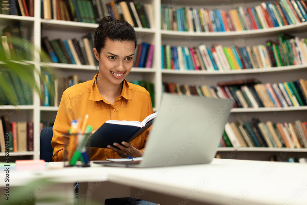 Virtual lecture. Happy mixed race woman sitting in library, holding book, having online class and looking at laptop