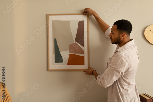 Young black man hanging painting, putting picture frame on wall indoors. Modern home decor and interior design