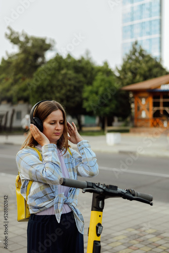 portrait of a young woman with an electric scooter in headphones and with a yellow backpack in the summer on a city street © sun_house_ann