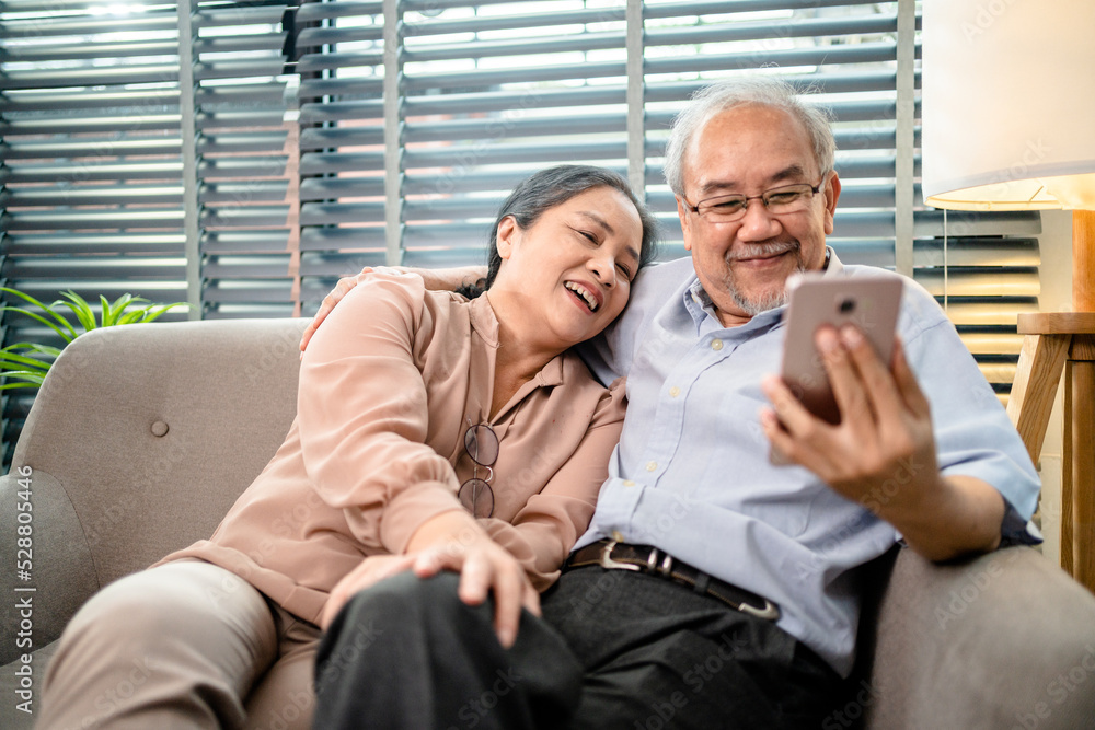 Happy Asian Elderly Couple sitting on couch while using smartphone video call online together in living room, holiday activities for retired couples, Senior health care and wellness mental concept.