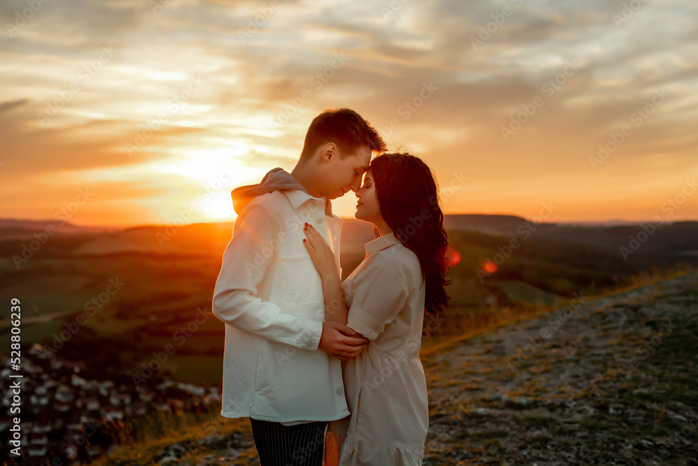A couple in love, a guy and a girl are standing on a mountain at sunset hugging and kissing
