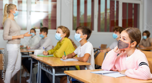 School girl in face mask sitting at desk in classroom on background with classmates and teacher