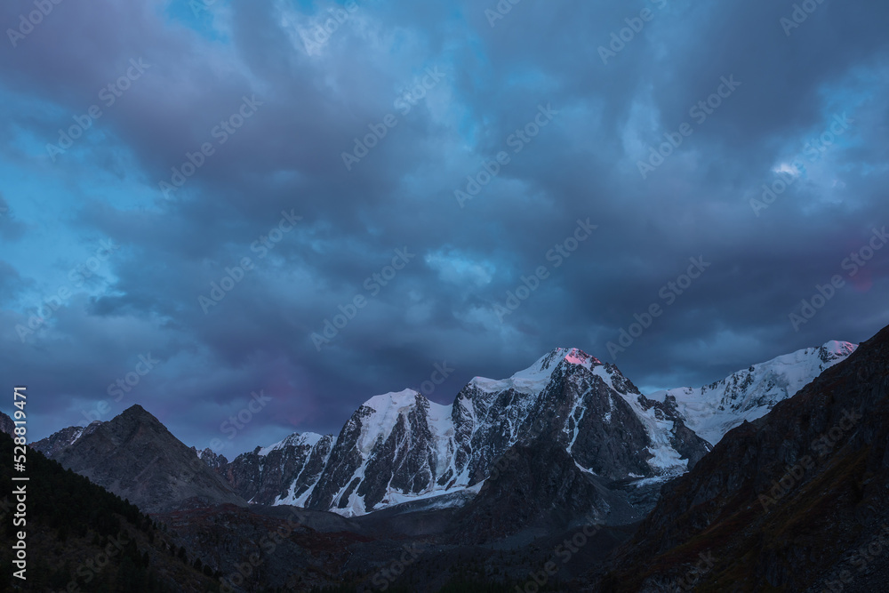 Atmospheric landscape with sunset pink reflection on huge snowy mountain top in dramatic sky. Scenic view to giant snow mountains in dusk. High snow-covered mountain range silhouettes in twilight.