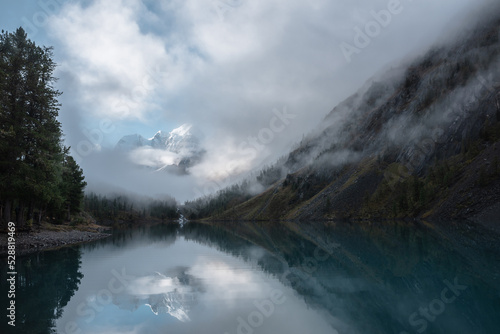Tranquil scenery with snow castle in clouds. Mountain creek flows from forest hills into glacial lake. Snowy mountains in fog clearance, small river and coniferous trees reflected in calm alpine lake. © Daniil