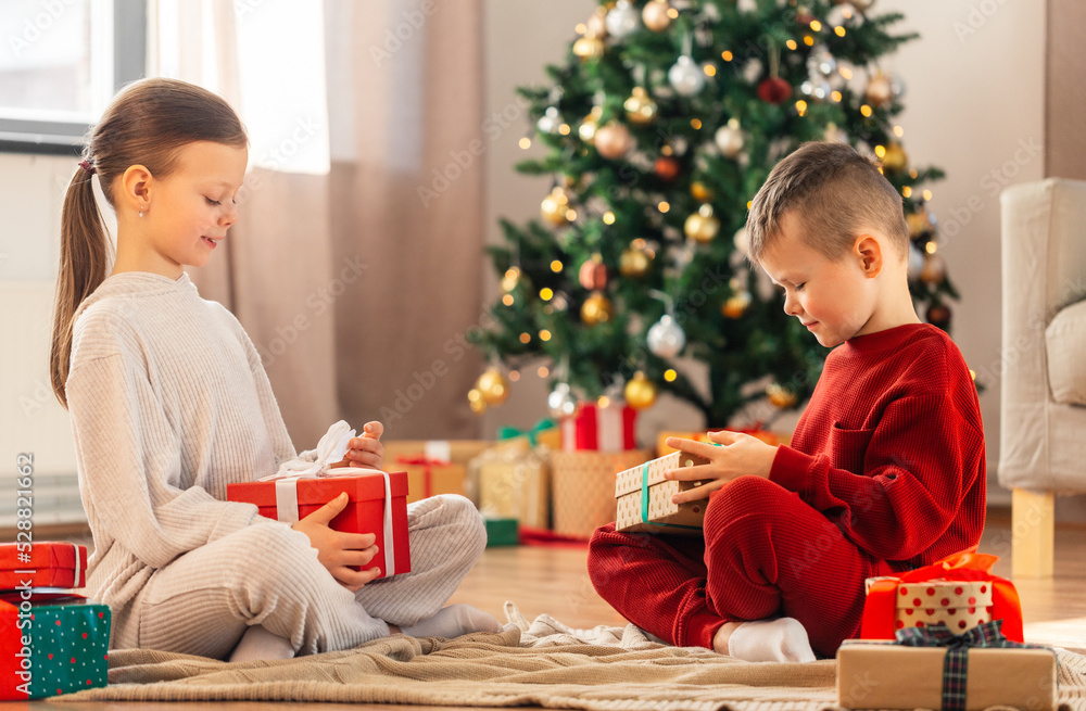 christmas, winter holidays and childhood concept - happy girl and boy in pajamas opening gifts sitting on floor in front of each other at home christmas, winter holidays and childhood concept - happy girl and boy in pajamas opening gifts sitting on floor in front of each other at home