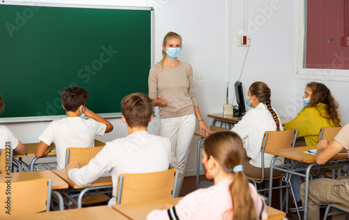 Woman teacher in face mask explaining new theme to children during class in primary school indoors