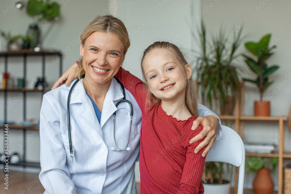 Portrait of friendly female doctor embracing with girl patient in hospital, looking and smiling togethe at camera Portrait of friendly female doctor embracing with girl patient in hospital, looking and smiling togethe at camera