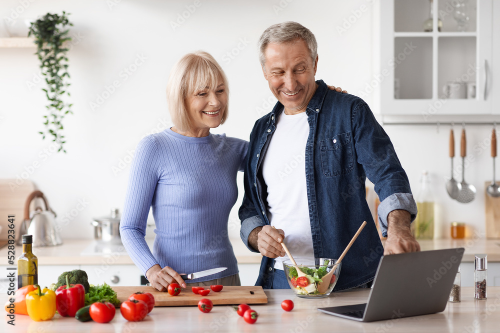 Loving senior spouses cooking together at home, using laptop Loving senior spouses cooking together at home, using laptop