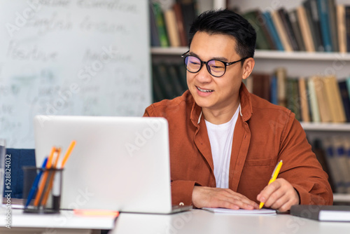 Japanese Male Tutor At Laptop Taking Notes Sitting At Workplace