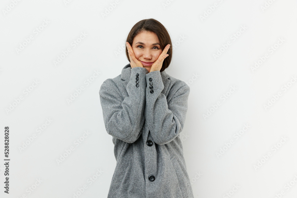 cute attractive middle aged woman is standing in a stylish long gray coat and smiling looking at the camera holding her hands near her face