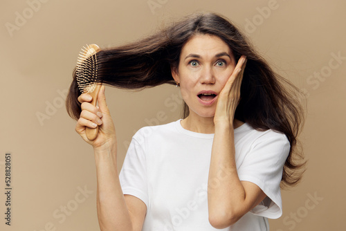 beautiful attractive middle-aged brunette woman combing her long hair with a wooden comb while standing in a white poppy and looking very surprised at the camera while standing on a beige background