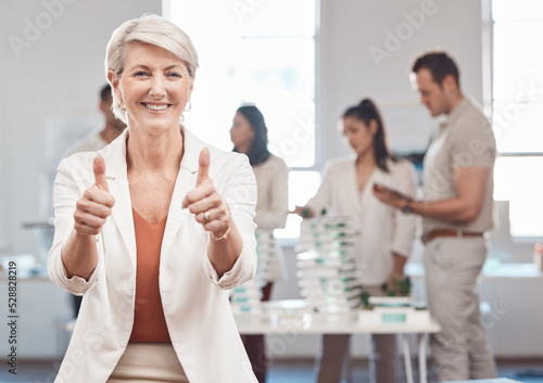 Thumbs up, motivation and yes with a woman leader, manager or CEO working in an office with her team in the background. Thank you, winner and success with a mature female business woman at work