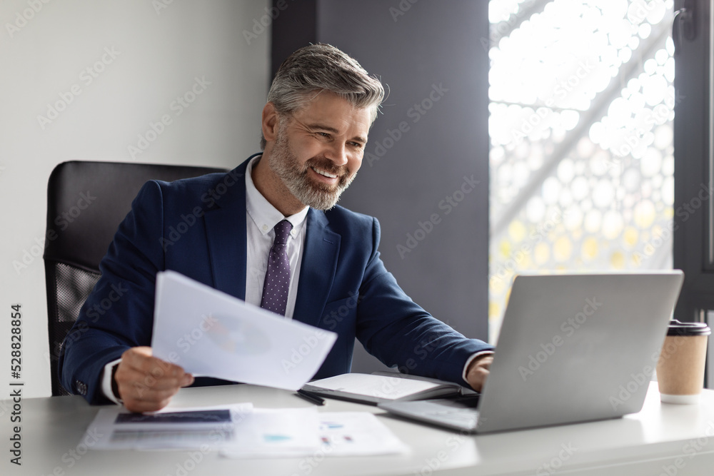 Successful middle aged entrepreneur working with laptop and papers in modern office Successful middle aged entrepreneur working with laptop and papers in modern office