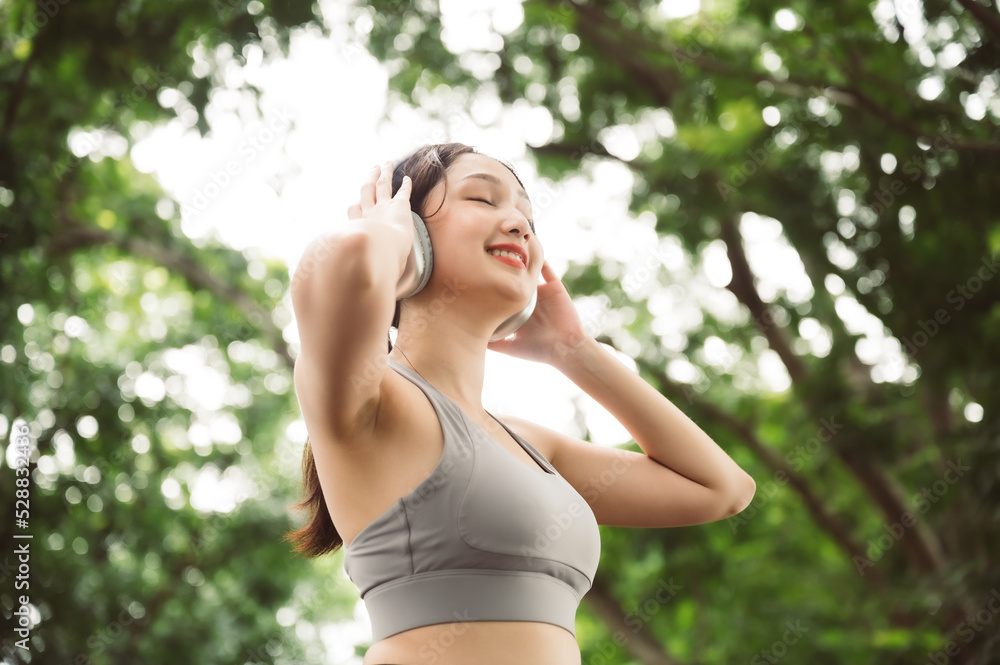 Asian woman trainer is engaged in fitness in public park. Female wearing sport bra and using headphone listening to music. Healthy lifestyle, sports outdoor activities in park
