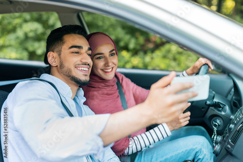 Cheerful glad millennial arab man and woman in hijab enjoy journey in new car and take selfie on smartphone