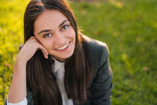 Close-up portrait of a freelancer young woman sitting on lawn in park resting from work in park. Coffee break. Positive person. Smiling and relaxing in summer