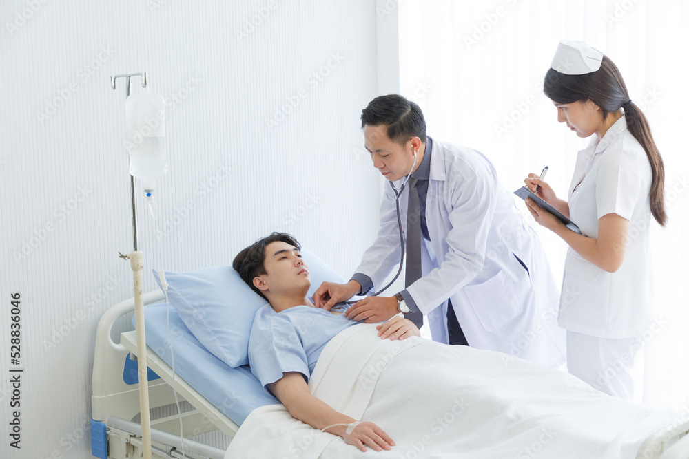 Senior doctor and young male patient who lie on the bed while checking symptom, consult and explain with nurse taking note and supporting in hospital wards. Senior doctor and young male patient who lie on the bed while checking symptom, consult and explain with nurse taking note and supporting in hospital wards.